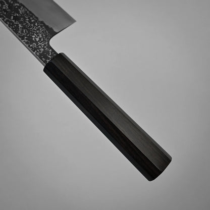 A close-up of a kurouchi damascus gyuto reveals a dark wooden handle and part of its textured metal blade against a plain grey background.