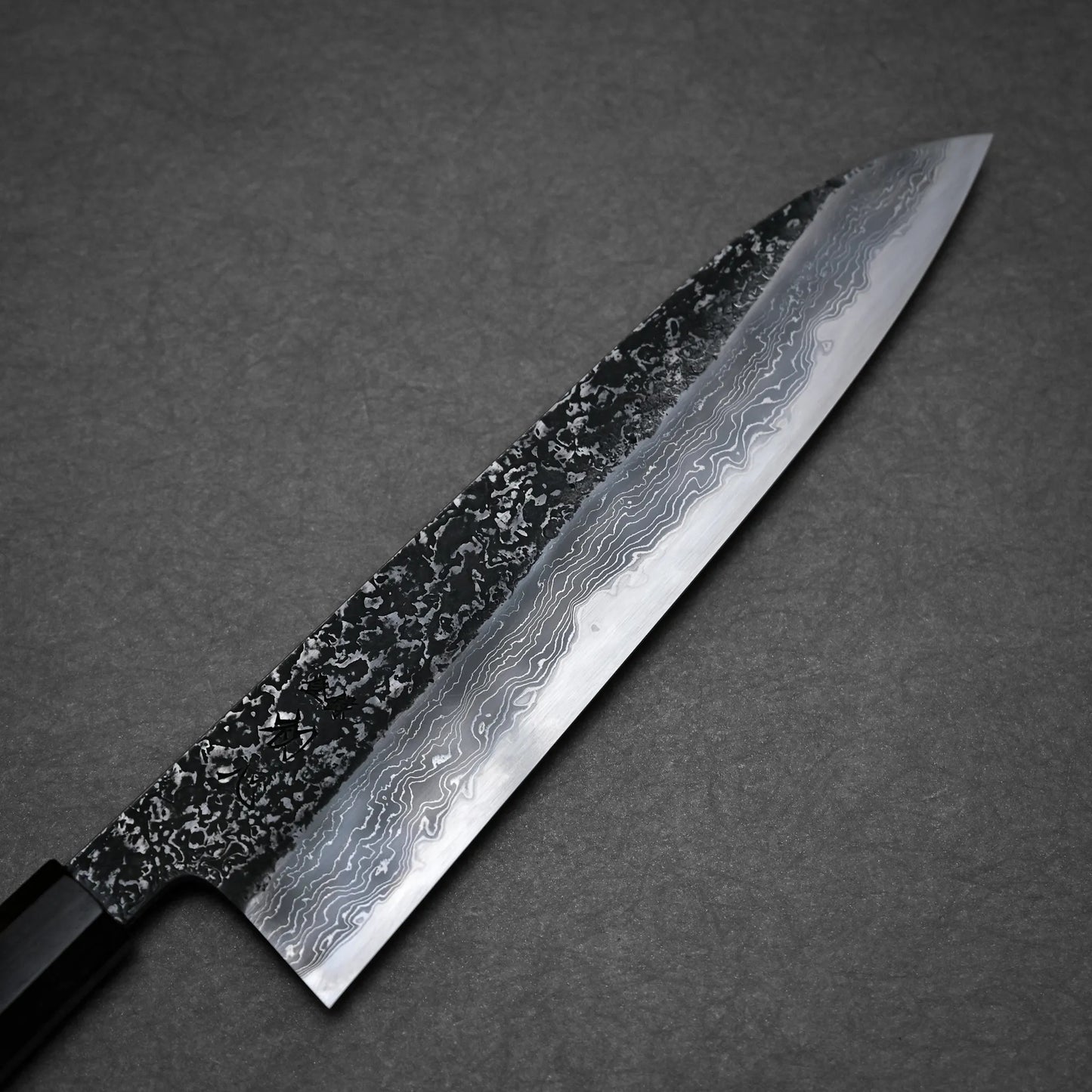 A close-up of a kurozome damascus Japanese chef's knife with an intricate wavy blade pattern, displayed against a dark, textured background.