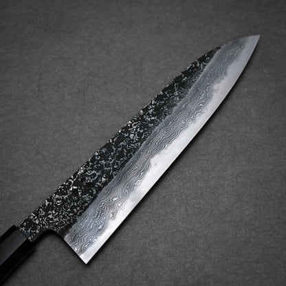 A close-up of a kurozome damascus Japanese chef's knife with an intricate wavy blade pattern, displayed against a dark, textured background.