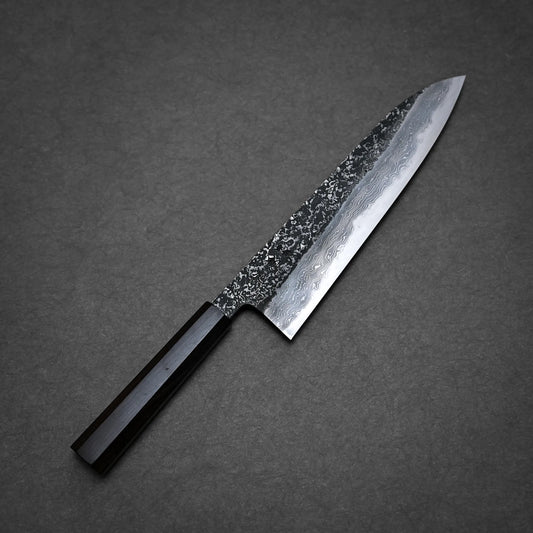 A Yoshihide Masuda kurozome damascus aogami#1 tall gyuto 240mm with a dark wooden handle and a patterned, polished steel blade rests on a dark textured surface. The blade features wavy patterns and a hammered finish near the spine, highlighting its artisanal kurouchi damascus gyuto design.