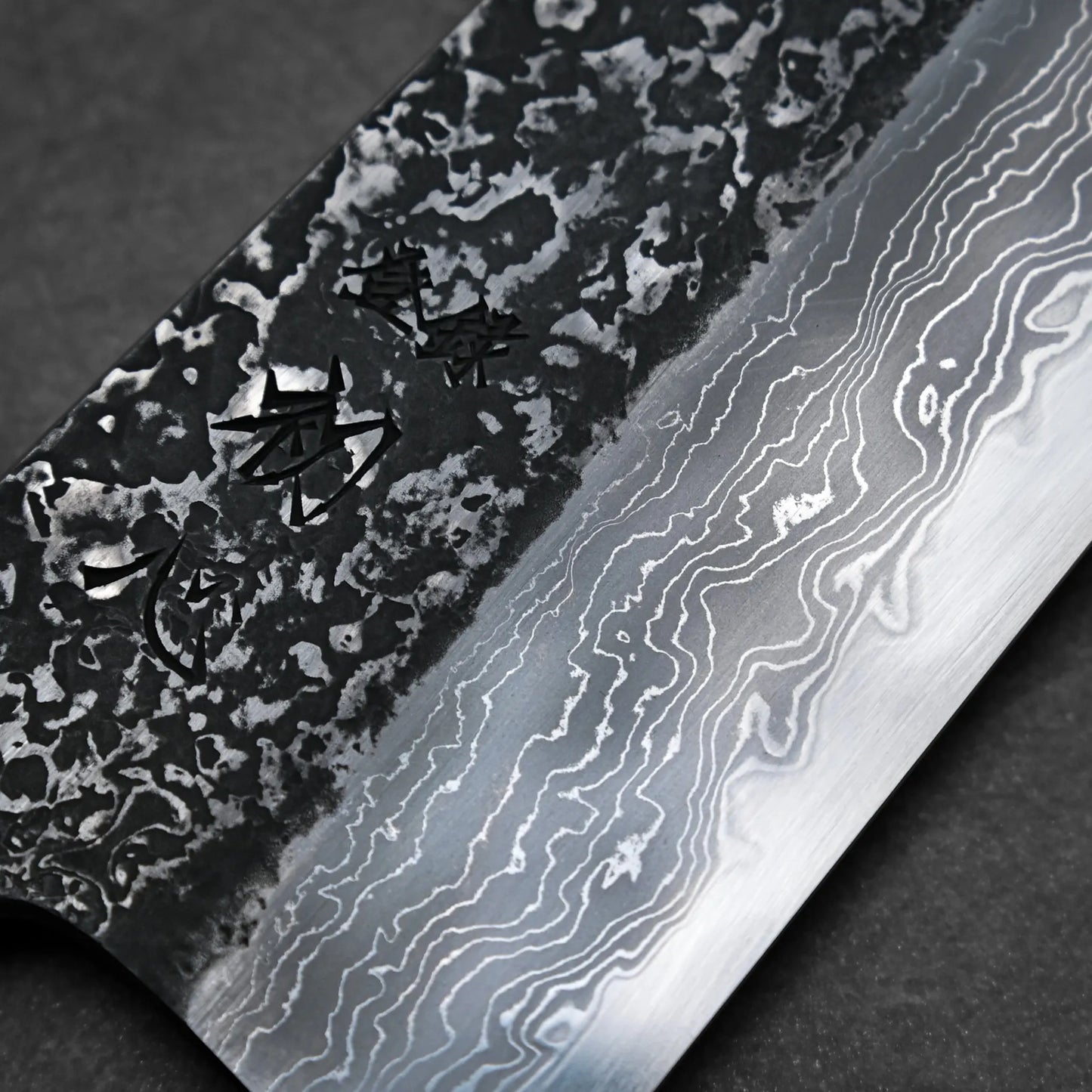 Close-up of a tall gyuto knife blade showing intricate Damascus steel patterns and engraved kanji characters on the textured upper part of the blade.