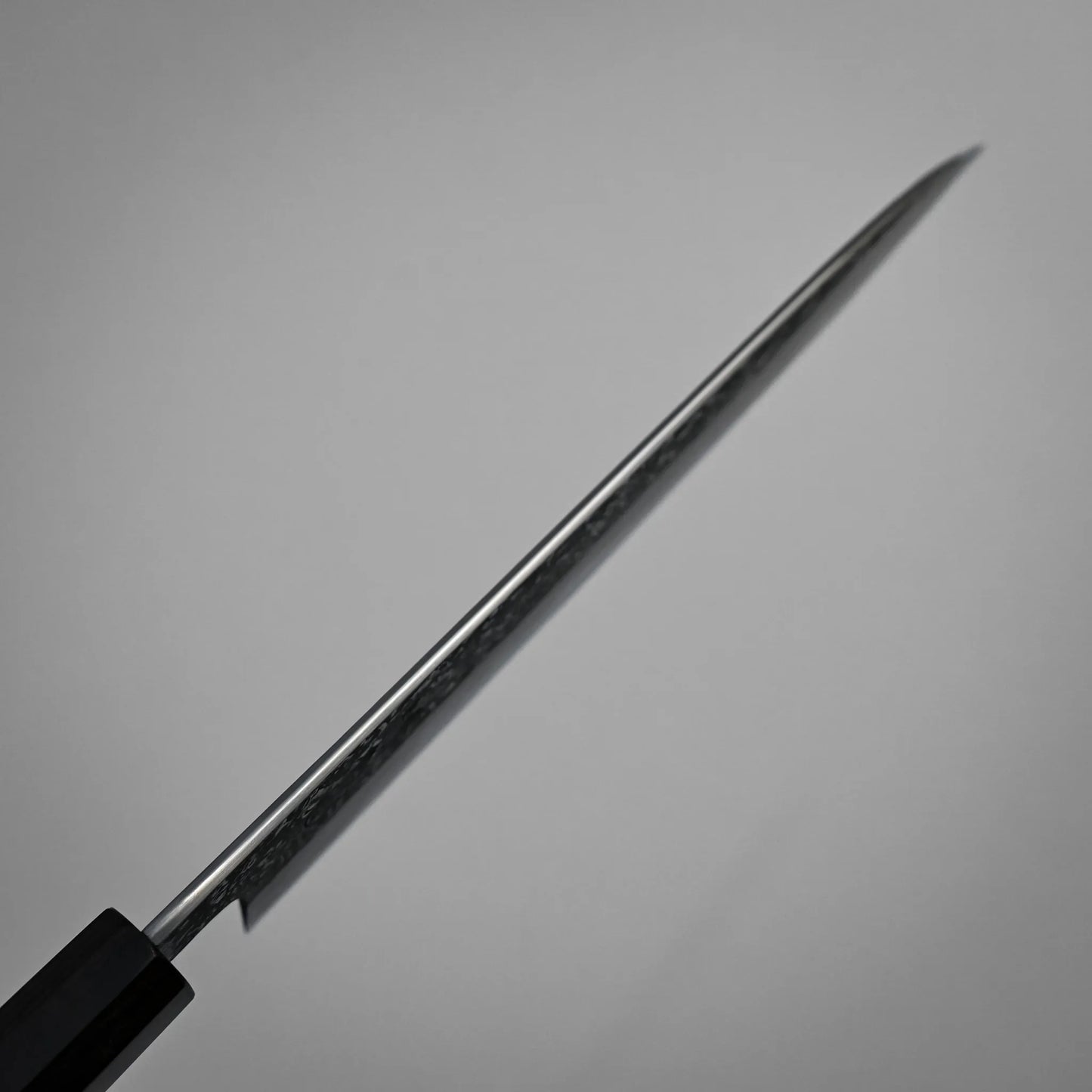 Spine shot of a kurozome damascus Japanese chef's knife blade angled diagonally against a plain, light gray background. The focus is on the blade’s edge, with the handle partially visible in the lower left corner.