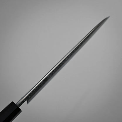 Spine shot of a kurozome damascus Japanese chef's knife blade angled diagonally against a plain, light gray background. The focus is on the blade’s edge, with the handle partially visible in the lower left corner.
