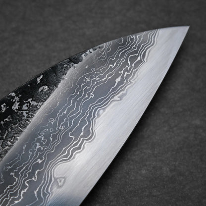 Close-up of a tall gyuto knife blade showing a wavy, layered pattern typical of Damascus steel on a dark gray background. The blade has a sharp edge and detailed texture.