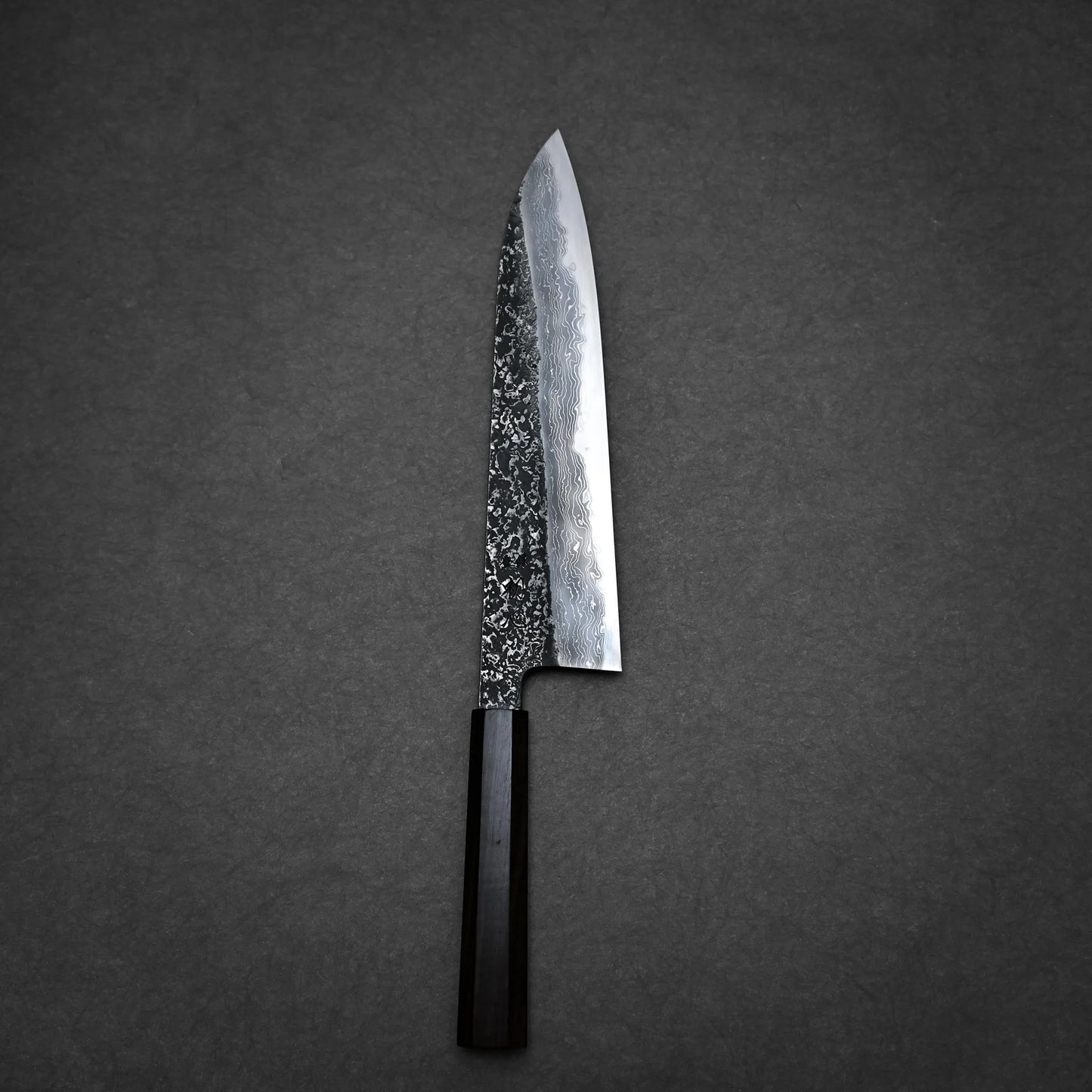 A tall gyuto knife with a dark wooden handle and a patterned, high-carbon steel blade rests on a dark, textured surface, echoing the elegance of Japanese craftsmanship.