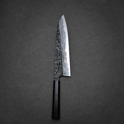 A tall gyuto knife with a dark wooden handle and a patterned, high-carbon steel blade rests on a dark, textured surface, echoing the elegance of Japanese craftsmanship.