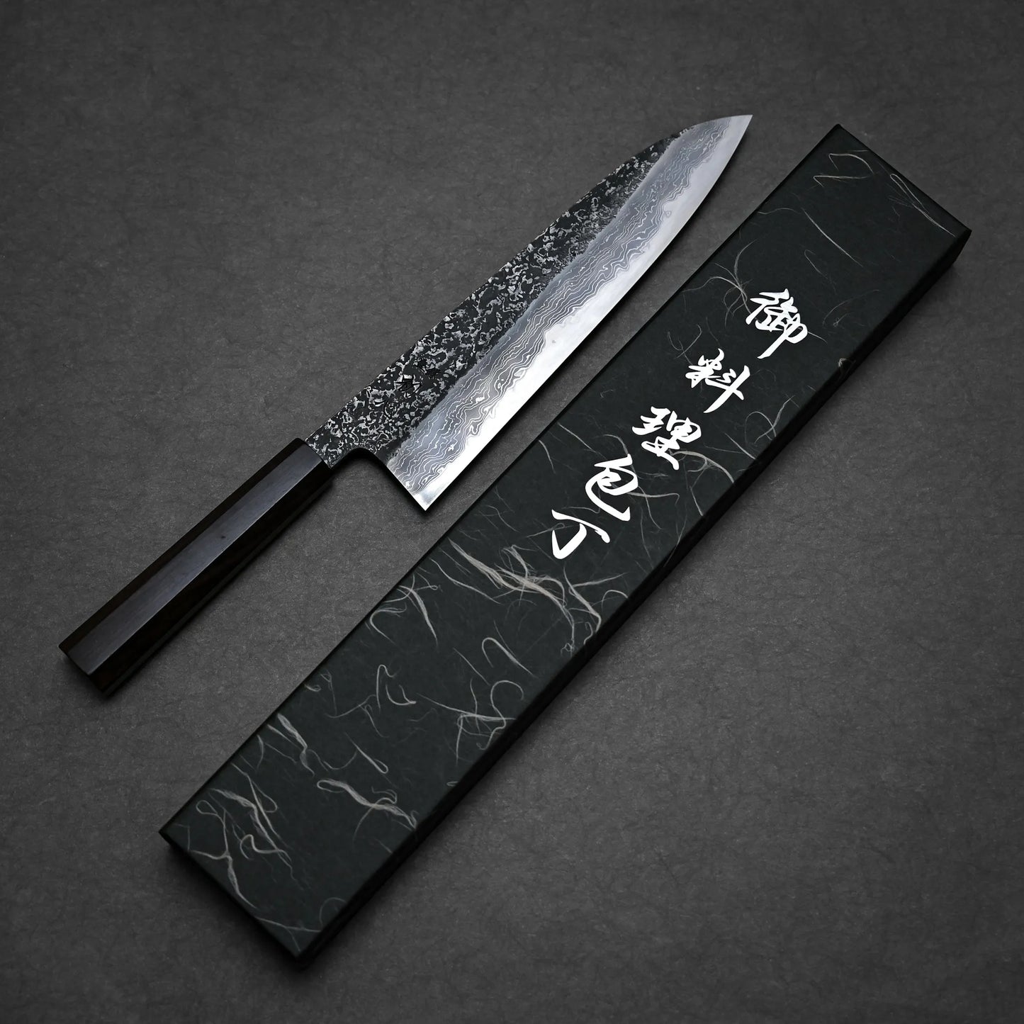 A kurouchi damascus gyuto with a hammered, patterned blade and a dark wooden handle is displayed next to a rectangular black box with white Japanese characters on the lid, set on a dark textured background.