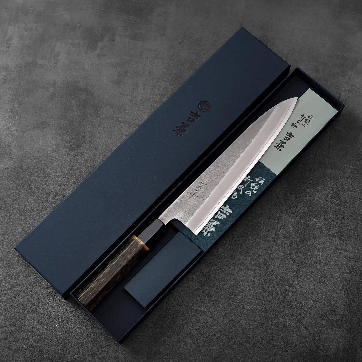 A gyuto with a burnt chestnut handle rests in a blue box, highlighting the contrast of materials and color.