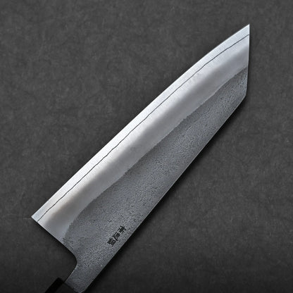 A close-up view of a bunka knife, a compact all-purpose Japanese knife, shows its polished blade with a visible hamon line resting on a dark textured background. The blade features a distinct straight edge and a textured lower half.