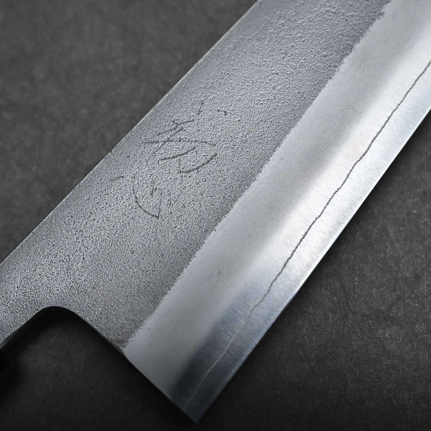 Close-up of a bunka knife blade with a visible hamon line and engraved Japanese characters, showing texture and detail of the metal against a dark background—an exquisite example of a compact all-purpose Japanese knife.