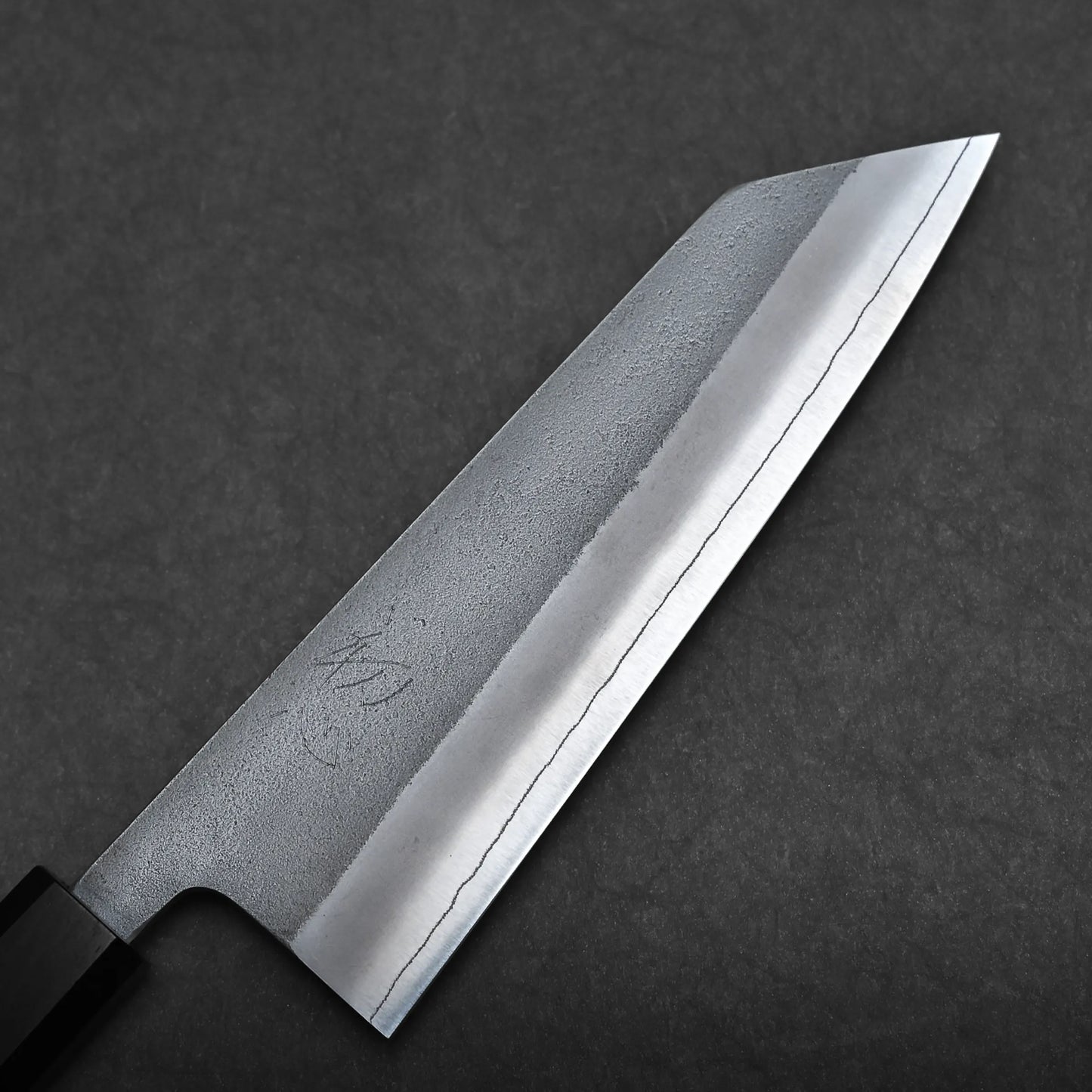 A close-up of a bunka knife—a compact all-purpose Japanese knife—with a straight edge and angled tip, showing a polished blade and subtle etched patterns on a dark surface background.