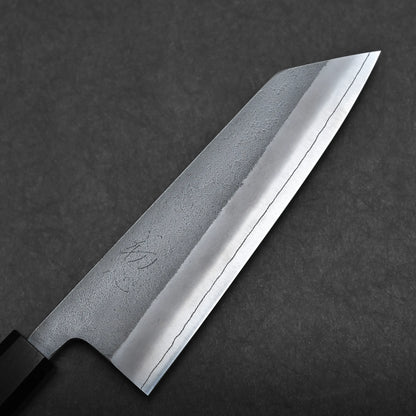 A close-up of a bunka knife—a compact all-purpose Japanese knife—with a straight edge and angled tip, showing a polished blade and subtle etched patterns on a dark surface background.