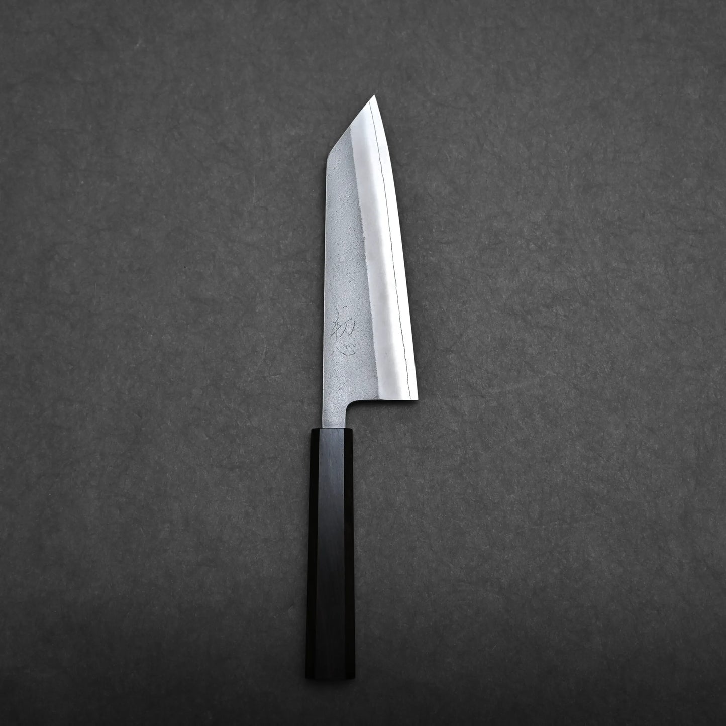 A compact all-purpose Japanese knife with a rectangular wooden handle and an angled, broad blade is placed on a dark grey textured surface, resembling the classic bunka knife style.