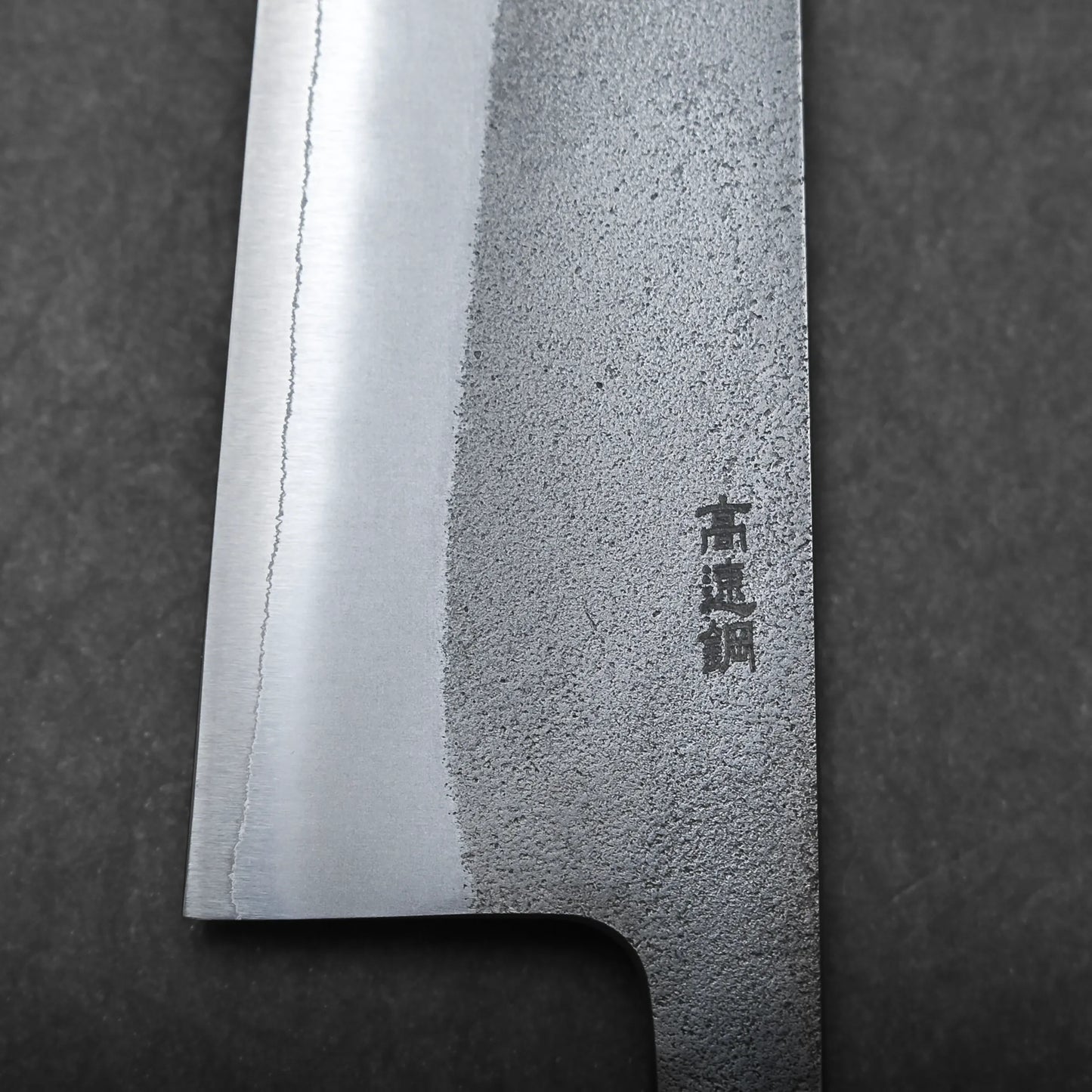 Close-up view of a Japanese chef's knife blade, featuring a textured pattern and engraved Kanji characters, set against a dark background.