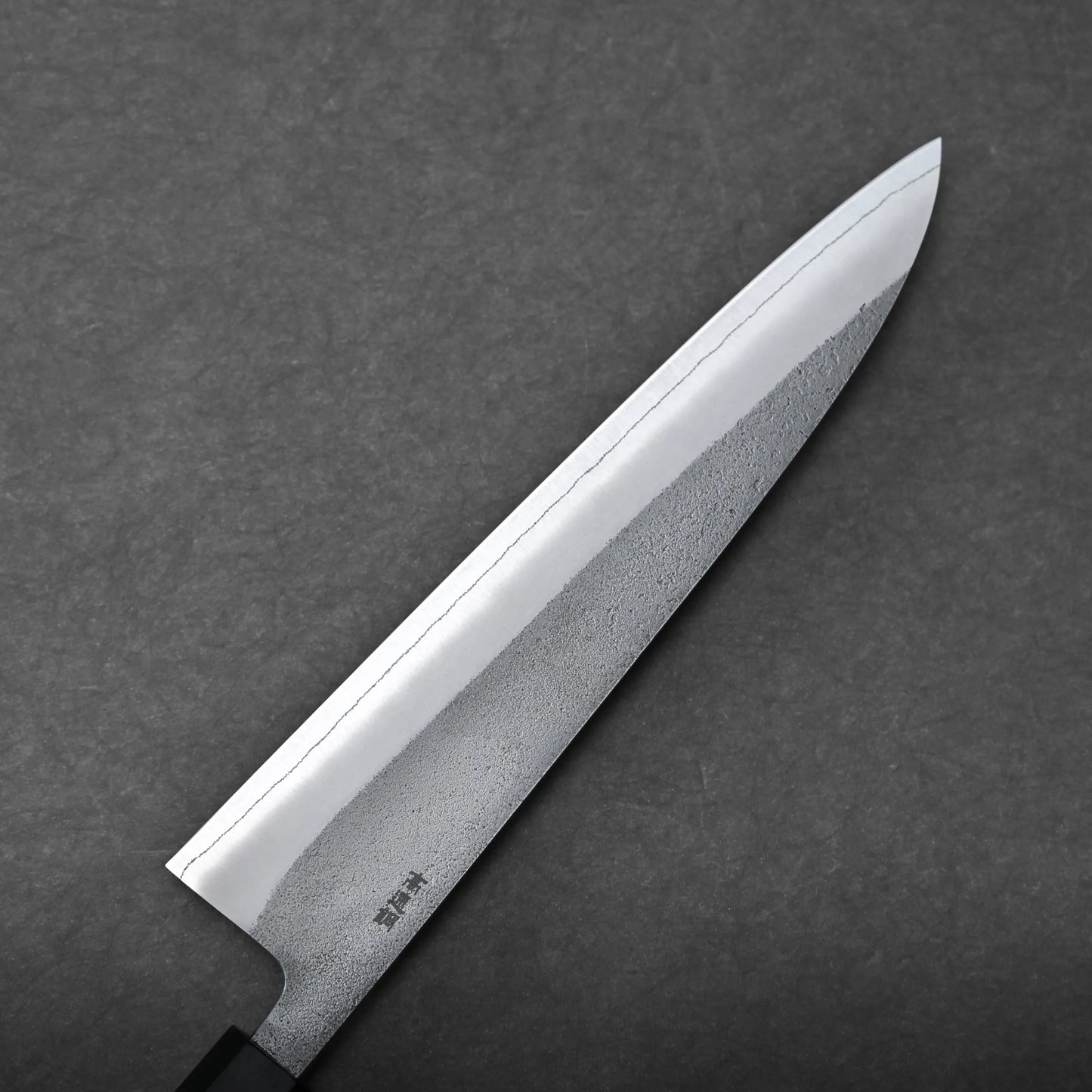 A close-up view of a Japanese chef's knife, or gyuto knife, with a sharp, shiny blade and a black handle, set against a dark gray textured background.