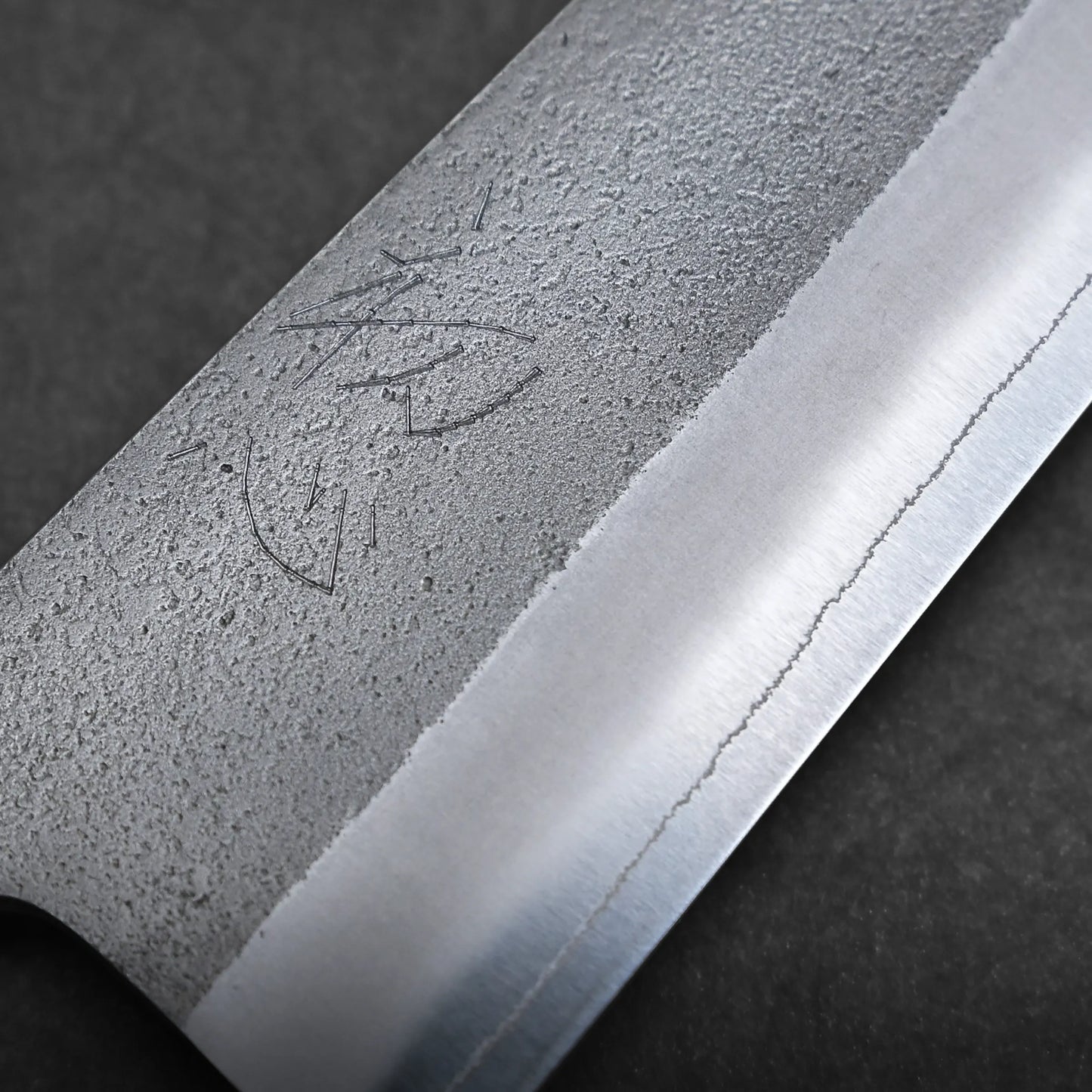 Close-up of a gyuto knife blade with a textured, matte finish on the upper part and a polished, sharp edge below. Engraved markings appear near the textured area. The background is dark and out of focus.
