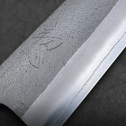 Close-up of a gyuto knife blade with a textured, matte finish on the upper part and a polished, sharp edge below. Engraved markings appear near the textured area. The background is dark and out of focus.