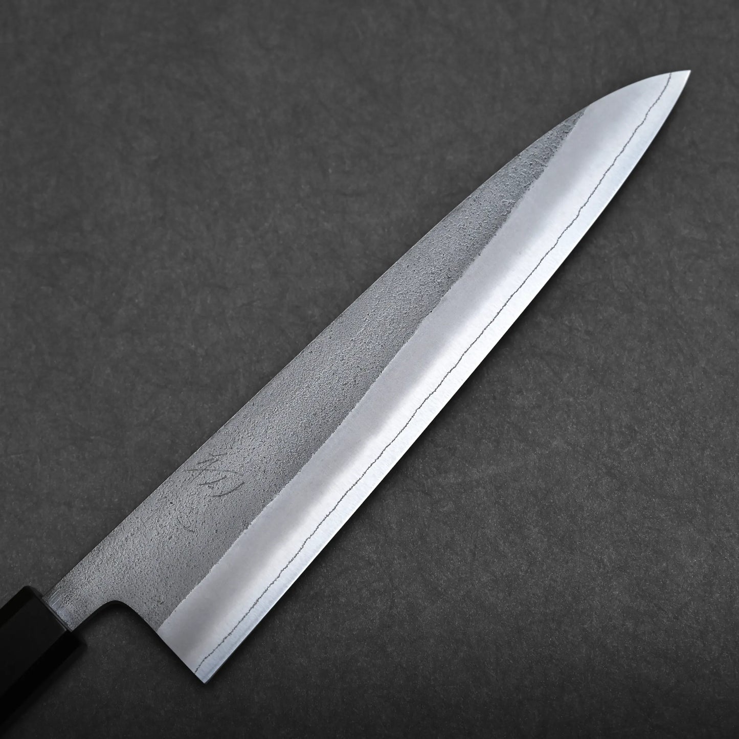 A close-up of a Japanese chef's knife with a sharp, polished blade and a dark handle, displayed against a textured dark gray background.