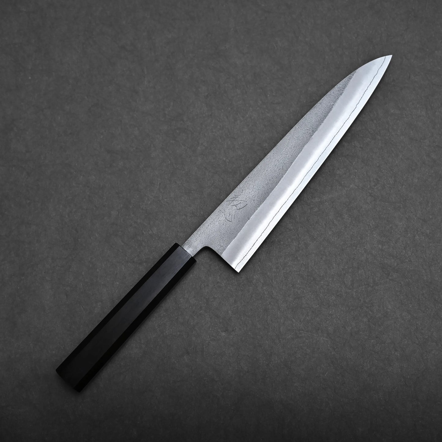 A Yoshikane nashiji SKD gyuto 210mm with a straight, sharp blade and a black, rectangular handle is placed on a dark gray, textured surface.