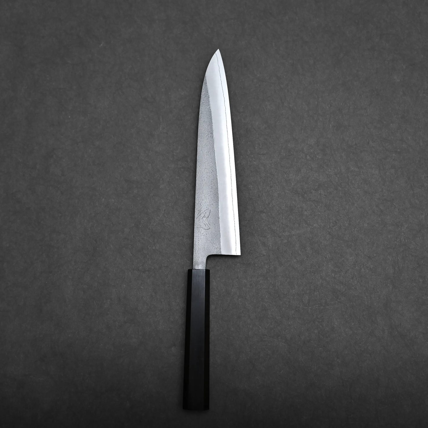 A single gyuto knife with a straight-edged blade and a black handle is placed on a dark gray surface, photographed from above.
