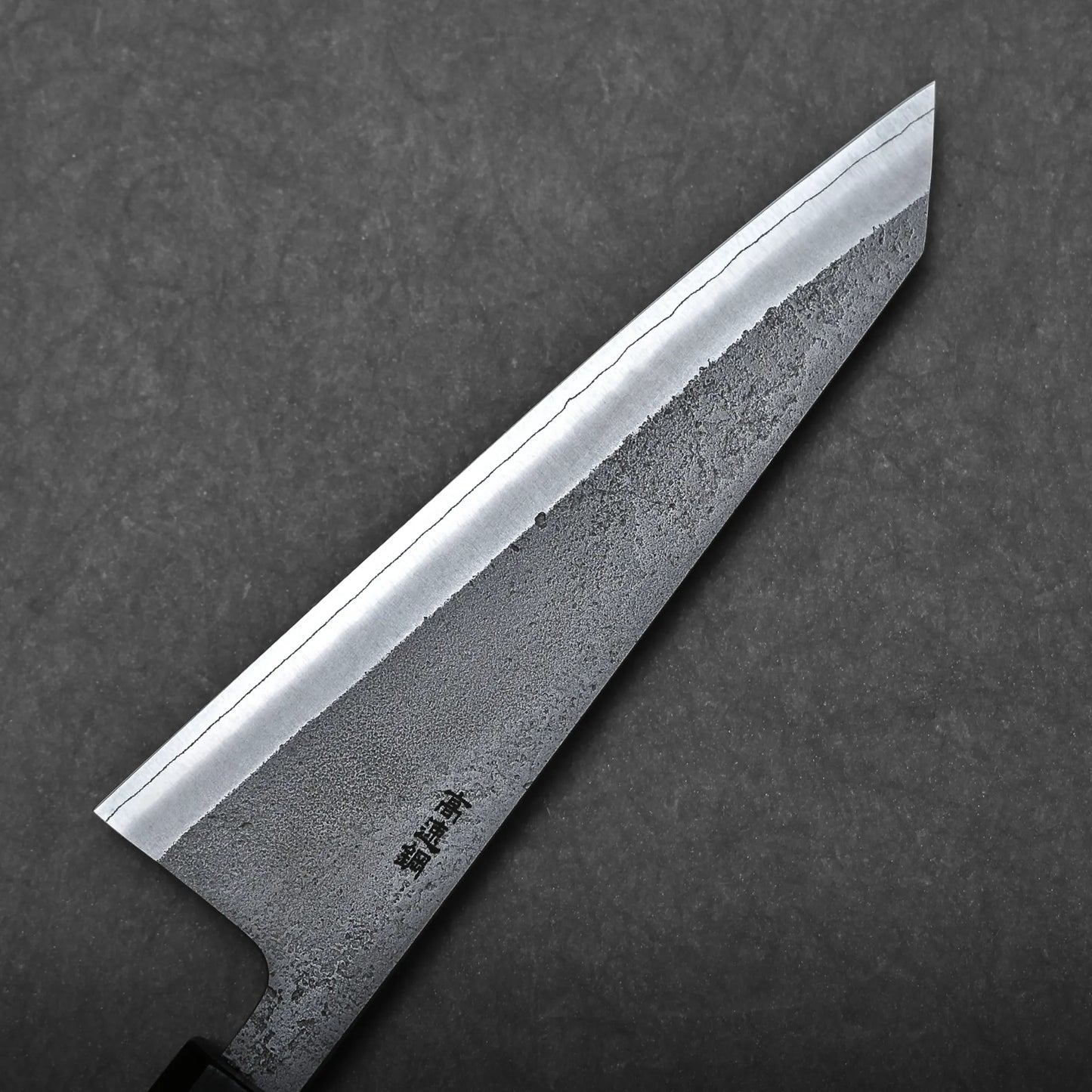 A close-up of a Japanese boning knife with a textured, patterned blade and sharp pointed tip, shown against a dark, solid background. Japanese characters are engraved near the honesuki knife’s handle.