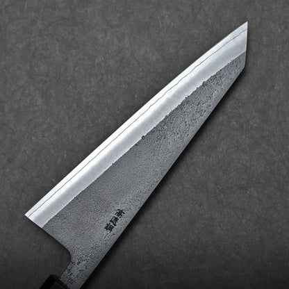 A close-up of a Japanese boning knife with a textured, patterned blade and sharp pointed tip, shown against a dark, solid background. Japanese characters are engraved near the honesuki knife’s handle.