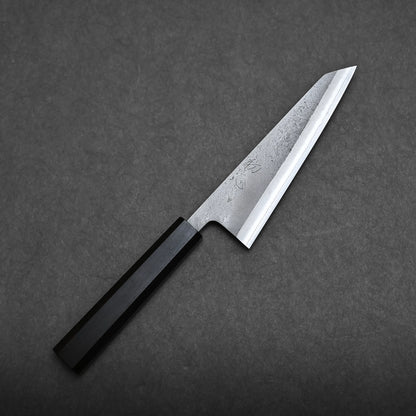 A Hatsukokoro x Yoshikane nashiji SKD honesuki 150mm with a wooden handle and a sharp, pointed blade lies diagonally on a dark, textured surface.