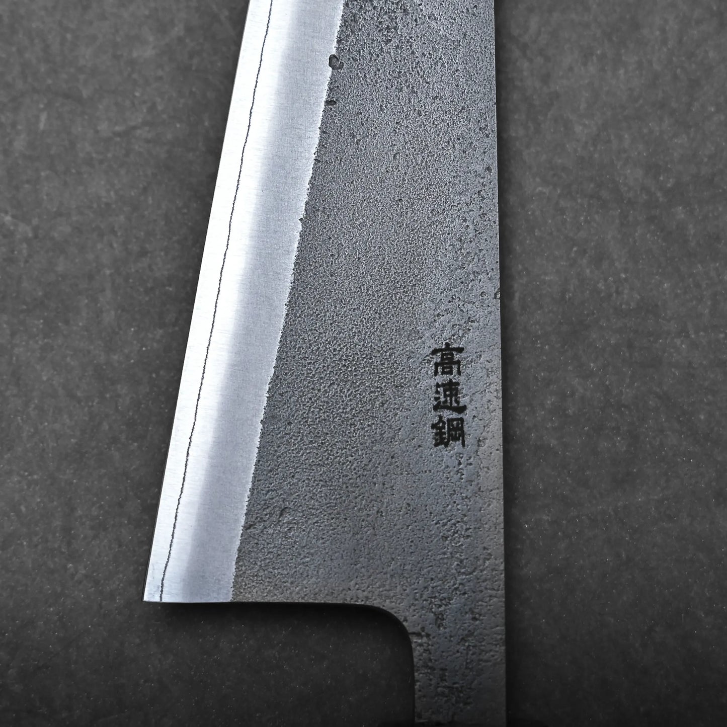 Close-up of a honesuki knife blade on a dark background, showing its sharp edge, textured steel surface, and black kanji characters engraved—highlighting the precision of this traditional Japanese boning knife.