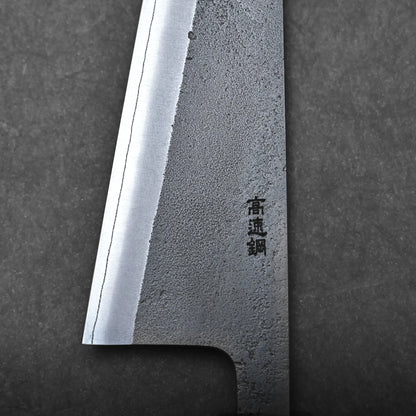 Close-up of a honesuki knife blade on a dark background, showing its sharp edge, textured steel surface, and black kanji characters engraved—highlighting the precision of this traditional Japanese boning knife.