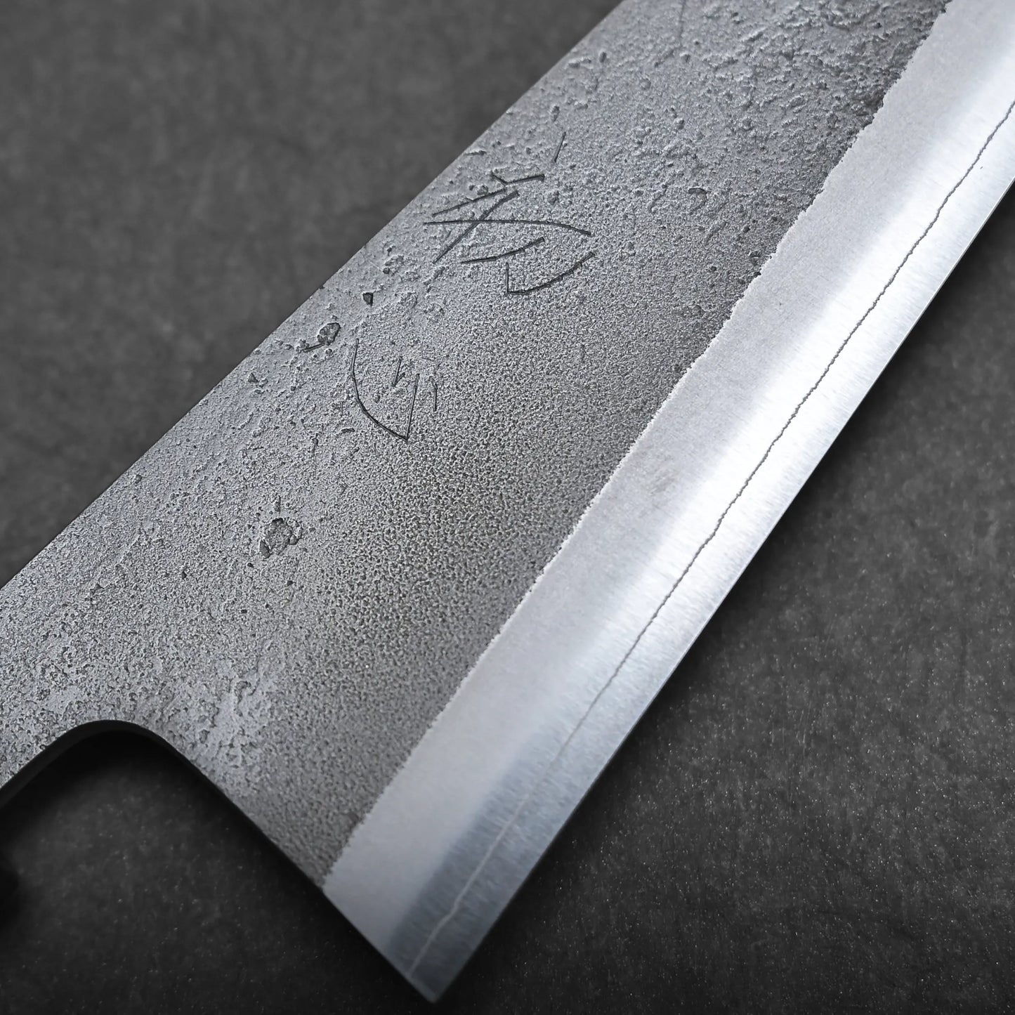 Close-up view of a honesuki knife blade with a textured, forged surface and a sharp polished edge. Japanese characters are engraved near the spine of this Japanese boning knife. The background is dark and stone-like.
