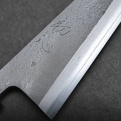Close-up view of a honesuki knife blade with a textured, forged surface and a sharp polished edge. Japanese characters are engraved near the spine of this Japanese boning knife. The background is dark and stone-like.