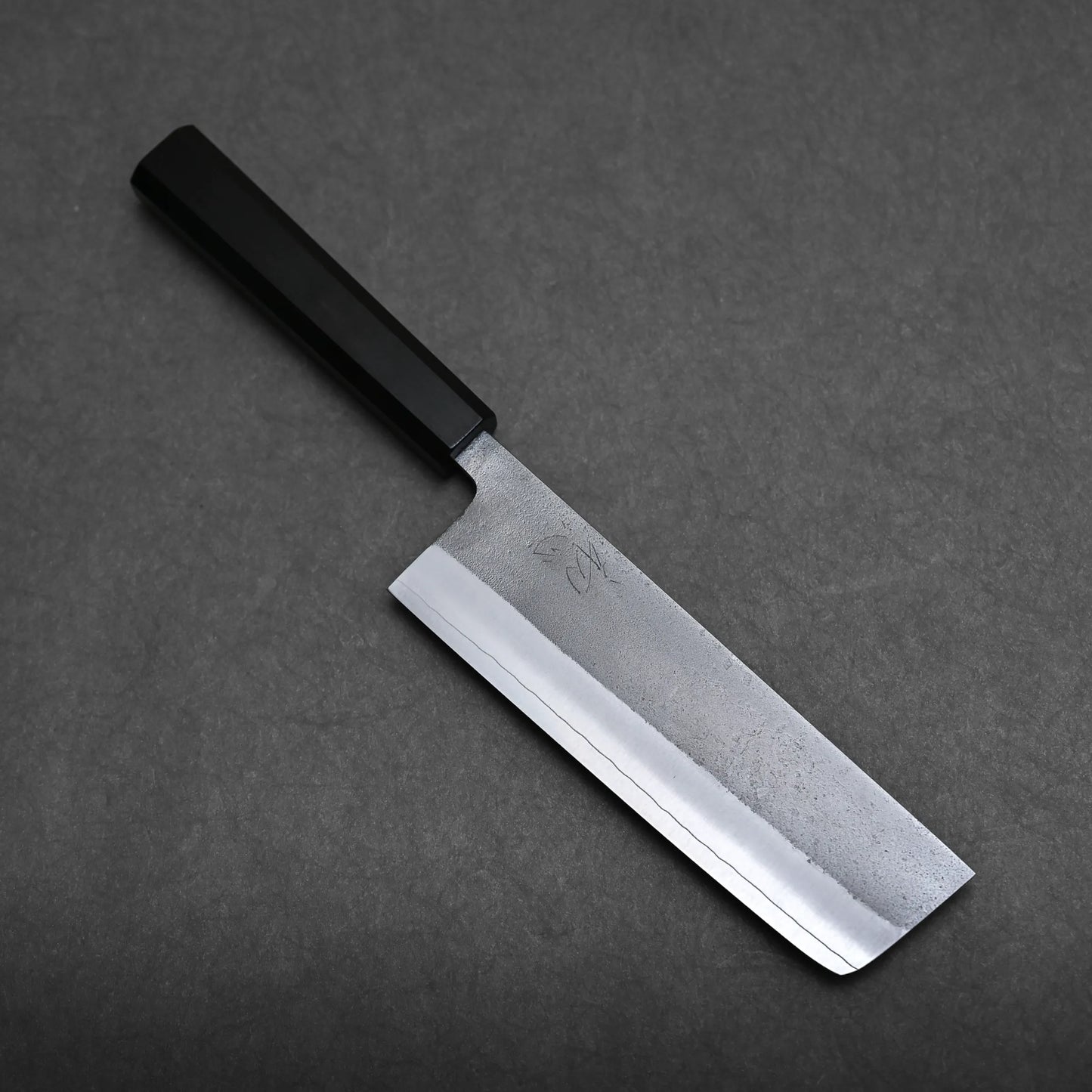 A Japanese-style kitchen knife with a black handle and a rectangular, straight-edged, steel blade is placed on a dark grey surface.