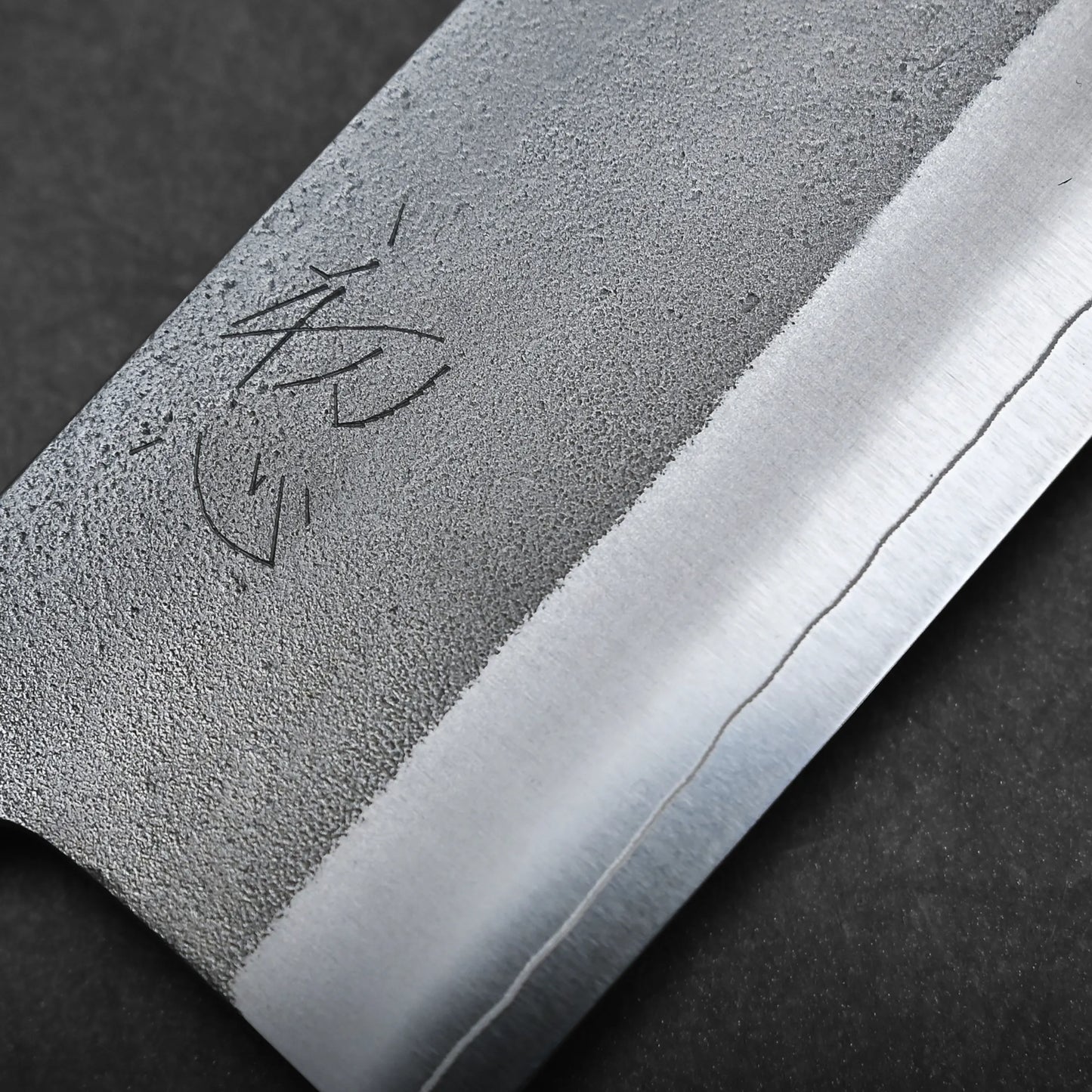 Close-up of a kitchen knife blade showing a textured surface, a polished edge, a faint wavy line near the edge, and an engraved symbol or writing on the upper part of the blade.