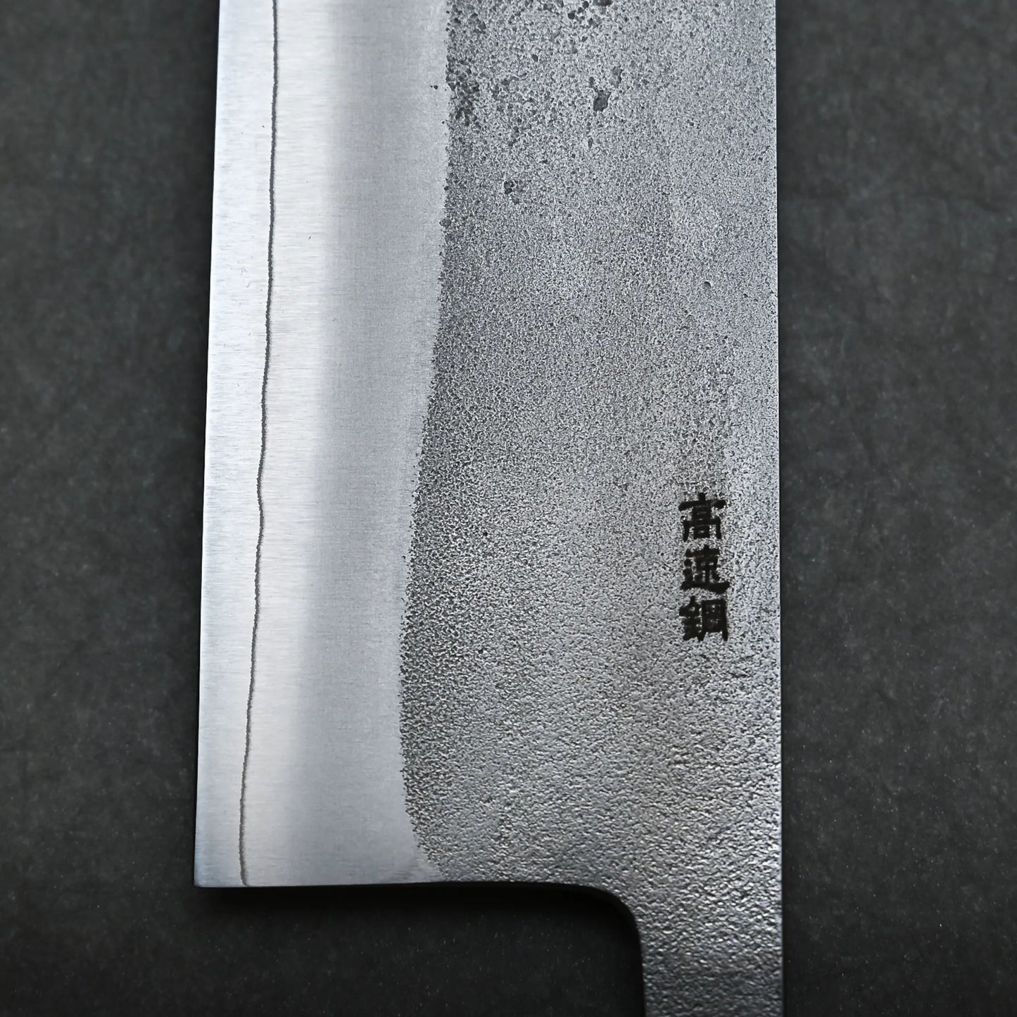 A close-up of a Japanese kitchen knife blade with visible patterns on the metal and Japanese characters engraved near the handle, photographed against a dark background.