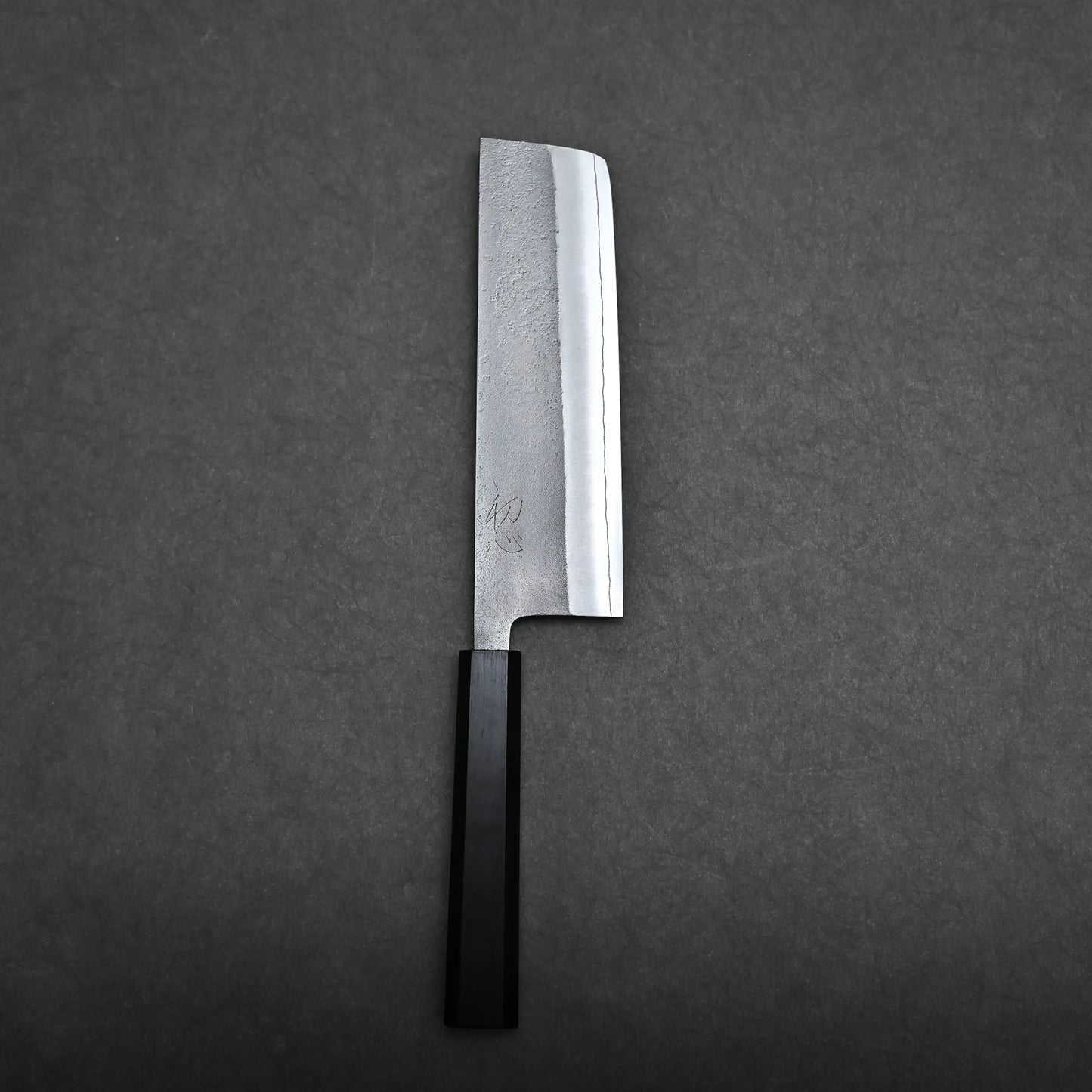 A Japanese-style kitchen knife with a rectangular blade and a black handle is placed on a dark gray textured surface. The blade has a slightly reflective finish and some visible markings near the handle.