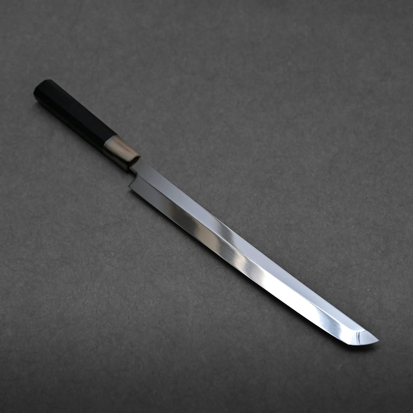 A long, single-edged honyaki sushi knife with a shiny, straight blade and a black handle is displayed on a dark gray surface.