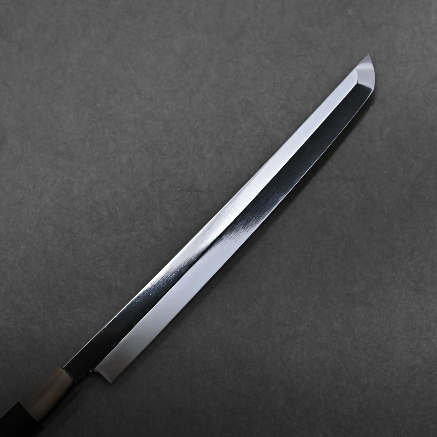A single-edged honyaki sushi knife with a shiny, reflective blade and sharp tip rests on a dark, textured surface. This mizuhonyaki aogami#1 sakimaru features a partially visible handle on the left side of the image.