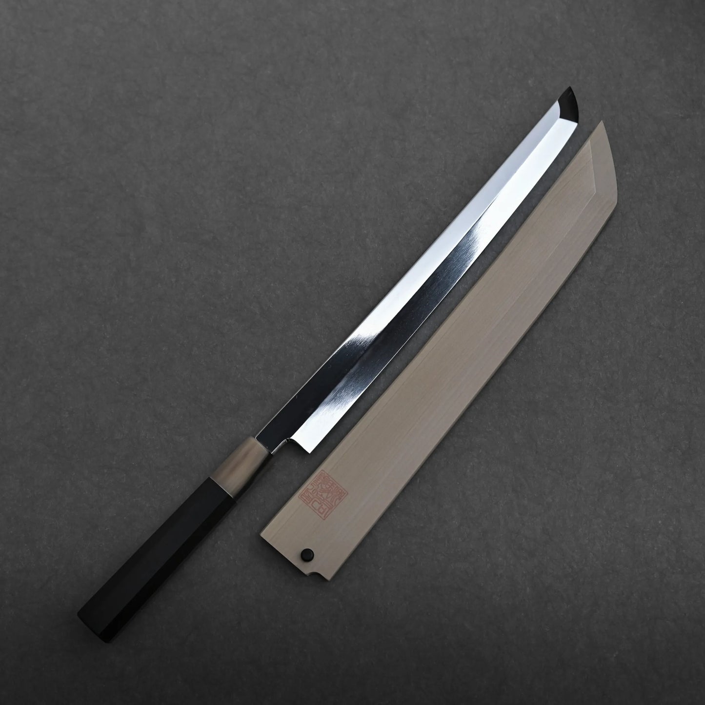 A Yoshikazu Ikeda aogami#1 honyaki sakimaru yanagiba 300mm with a black handle rests next to its wooden sheath on a dark gray surface. The mizuhonyaki aogami#1 sakimaru blade is shiny and straight, tapering to a sharp angled point.