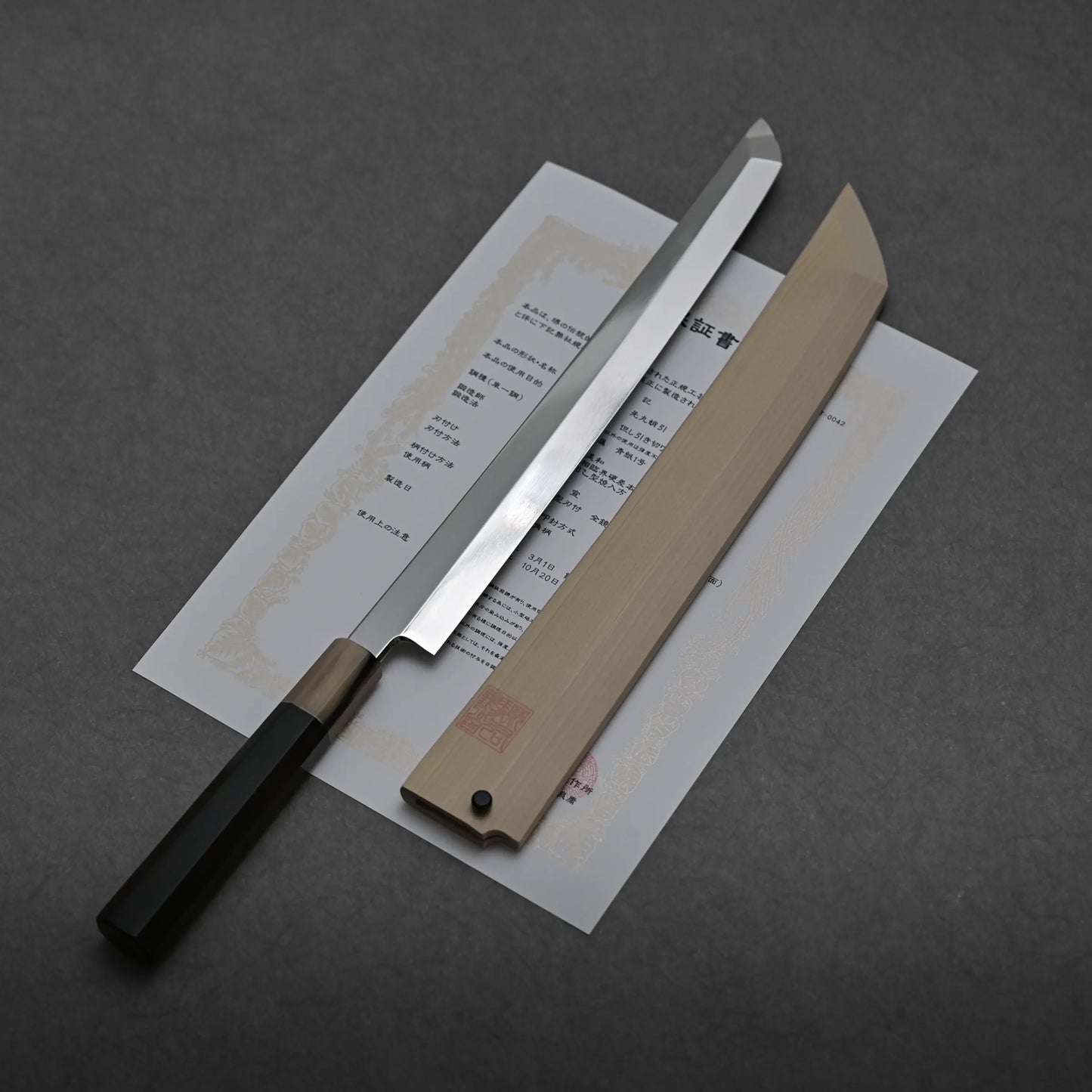 A mizuhonyaki aogami#1 sakimaru honyaki sushi knife with a wooden handle and sheath rests atop a certificate featuring Japanese text, set against a dark grey background.