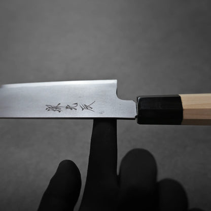 A gloved hand balances a Japanese petty knife on the tip of an index finger, with Japanese characters engraved on the blade against a plain gray background. It shows the balance point.