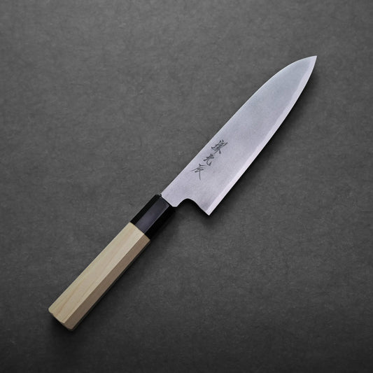 A Yoshikazu Ikeda shirogami#2 gyuto 180mm with a wooden handle and a shiny, sharp blade featuring engraved characters rests on a dark gray textured surface.
