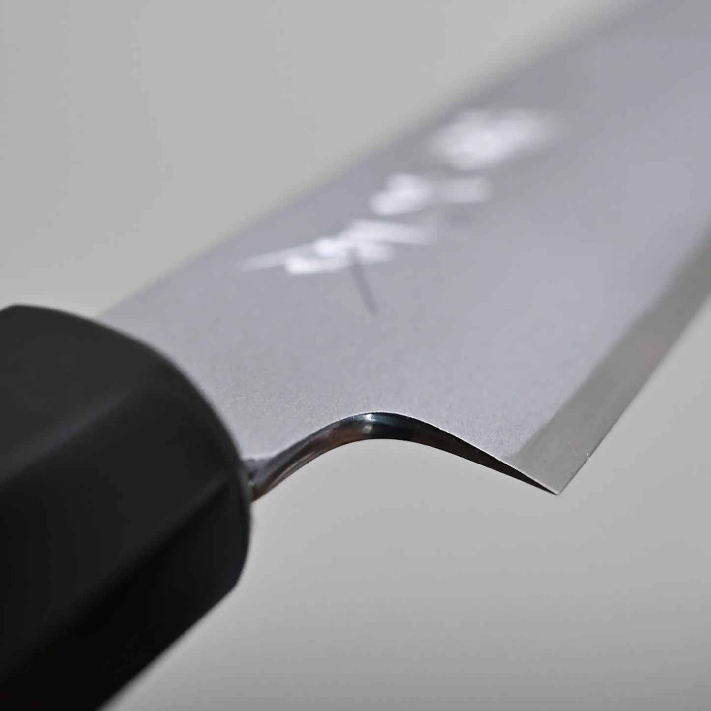 Close-up of the blade and handle of a petty knife, showing the choil finish and Japanese characters engraved on the metal. The background is plain and out of focus.