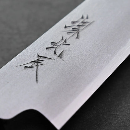Close-up of a Japanese petty knife blade featuring engraved Japanese characters on a smooth metallic surface with a sharp edge. The background is dark and slightly out of focus.