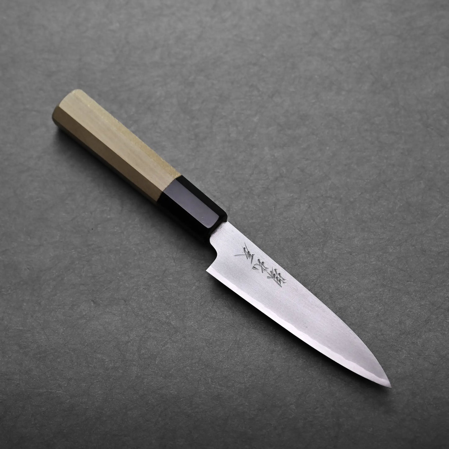 A small petty knife with a wooden handle and a shiny, pointed blade featuring Japanese characters rests on a dark, textured surface.