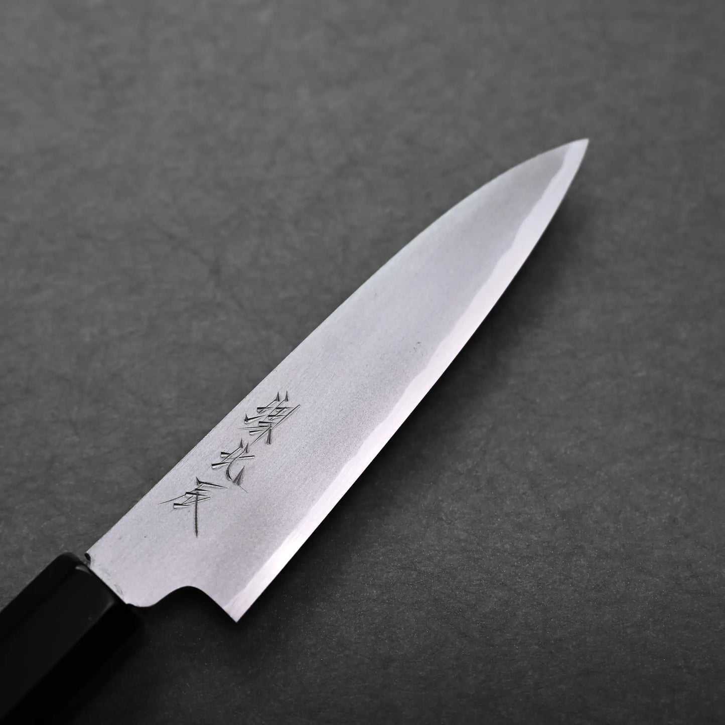 A close-up of a Japanese petty knife with a sharp, pointed blade and engraved characters on the metal, placed on a dark textured surface.
