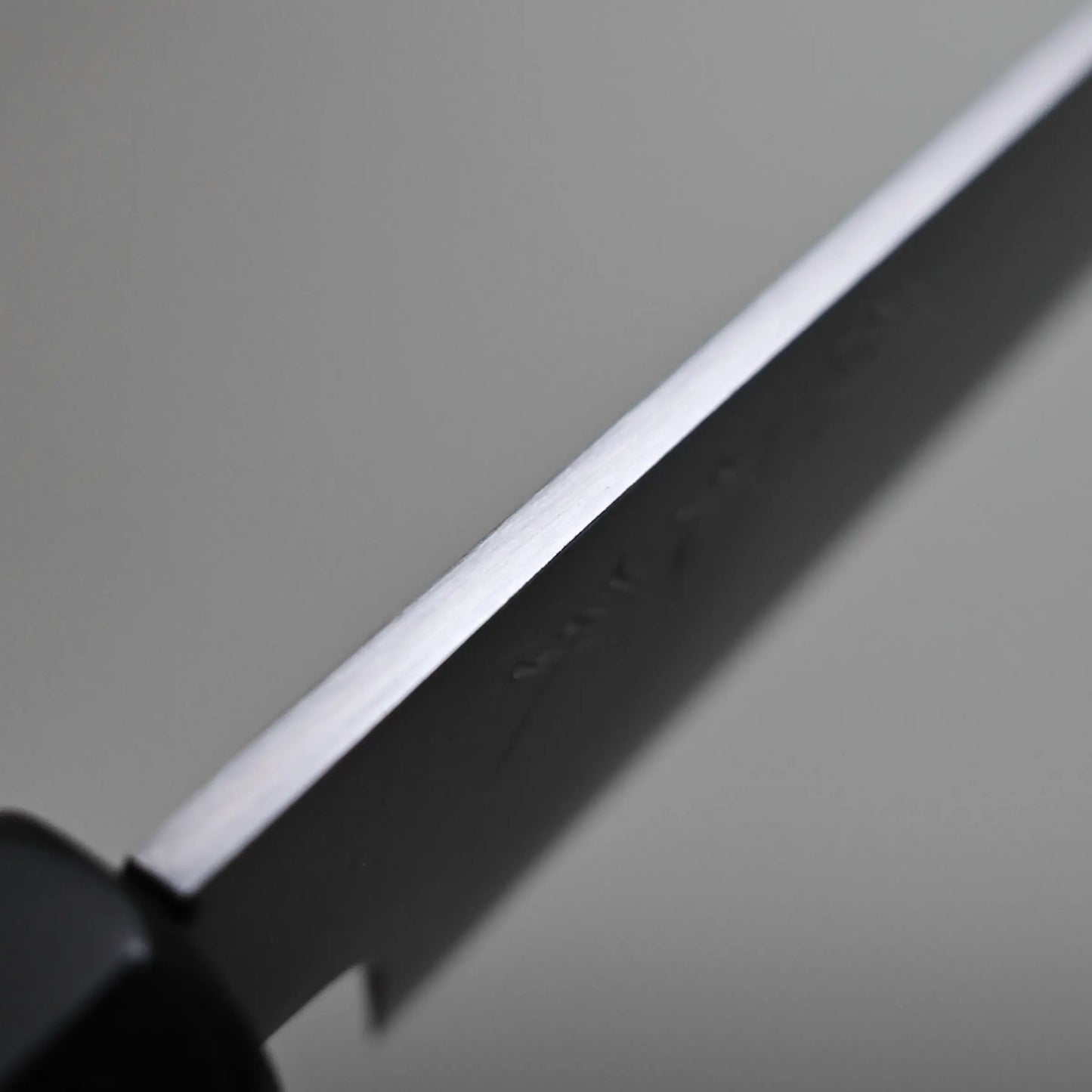 Close-up of a petty knife blade showing the spine and part of the black handle, set against a neutral gray background.