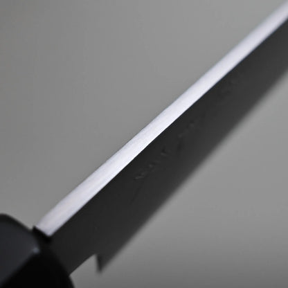 Close-up of a petty knife blade showing the spine and part of the black handle, set against a neutral gray background.