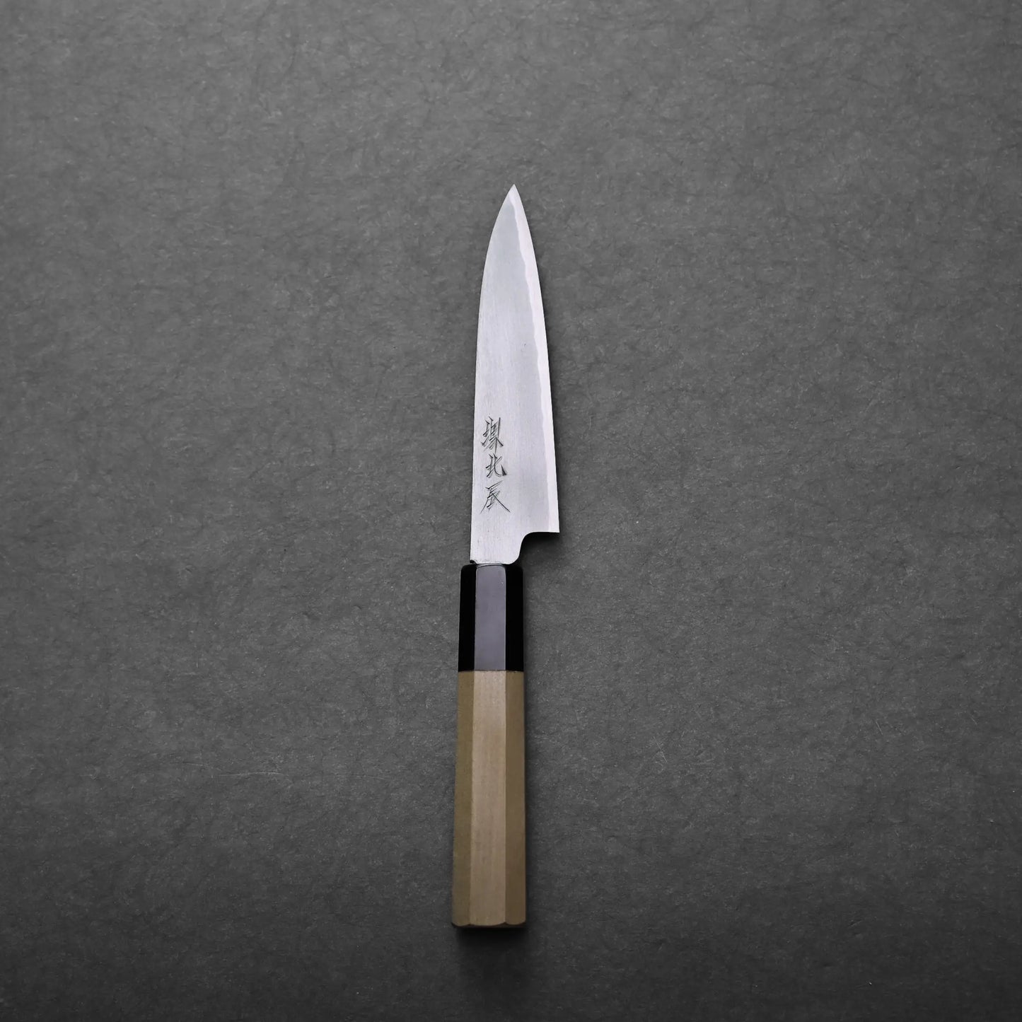 A single Japanese petty knife with a wooden handle and a pointed stainless steel blade, featuring Japanese characters on the blade, is placed on a dark gray textured surface.