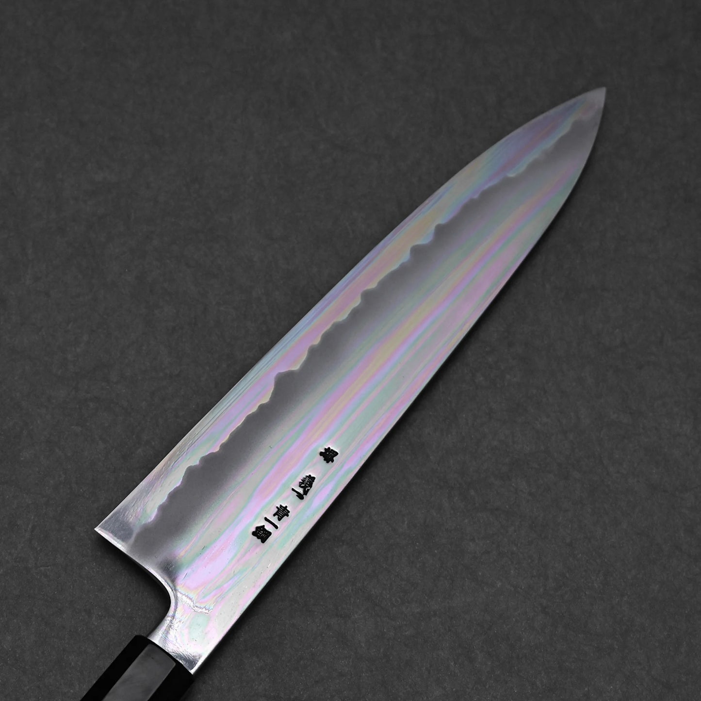 A close-up of a chef's knife shows its shiny, iridescent blade with subtle rainbow hues and visible kanji, placed on a dark textured background.