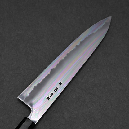 A close-up of a chef's knife shows its shiny, iridescent blade with subtle rainbow hues and visible kanji, placed on a dark textured background.